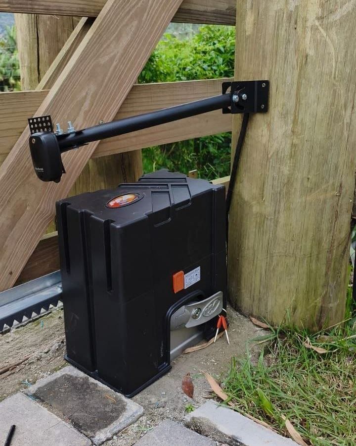 Motor de portão robusto, preto, instalado sobre base de concreto junto a um portão rústico de madeira, com cremalheira e fotocélula em braço metálico